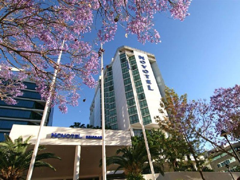 布里斯班阿莫拉酒店 外观 照片 The photo shows a modern hotel building, identified as "Novotel," rising above a landscaped area with palm trees. In the foreground, there are vibrant purple flowering trees, likely jacarandas, which add to the picturesque scene. The sky is clear and